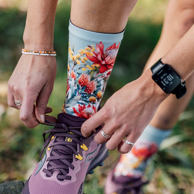 Person wearing colorful floral socks and a smartwatch, tying running shoes outdoors.